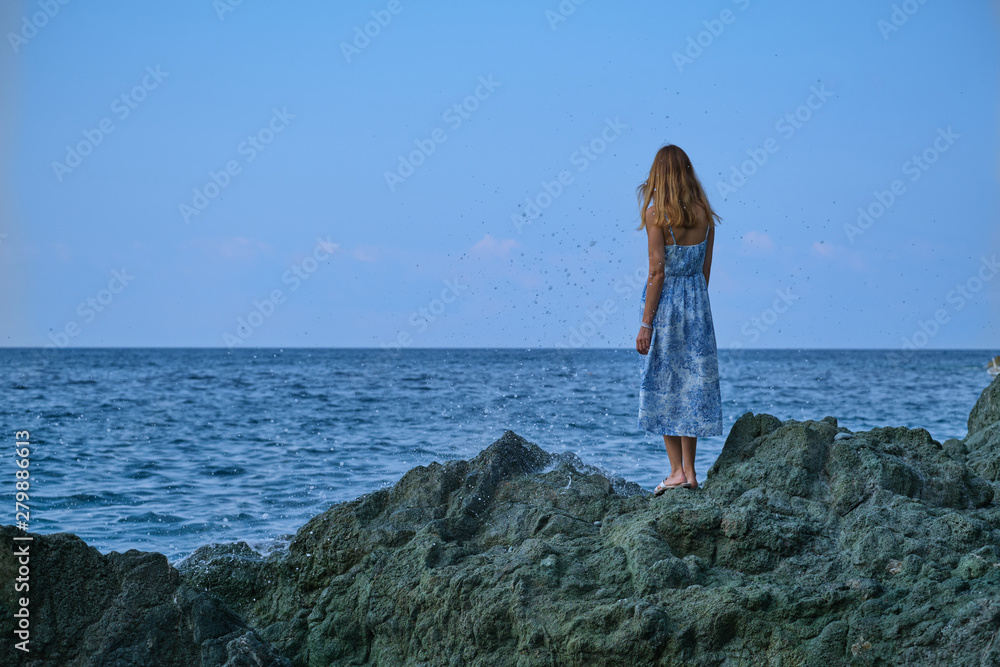 A beautiful young girl in a blue dress is standing on a rock and looking at the sea. A beautiful young girl in a blue dress is standing on a rock and looking at the sea.