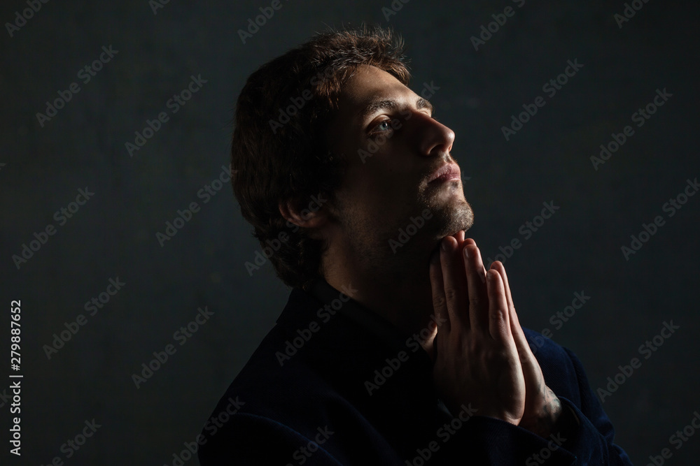 Portrait of a young pensive man on a dark background, looking to the side. Hands in prayer gesture. Portrait of a young pensive man on a dark background, looking to the side. Hands in prayer gesture.