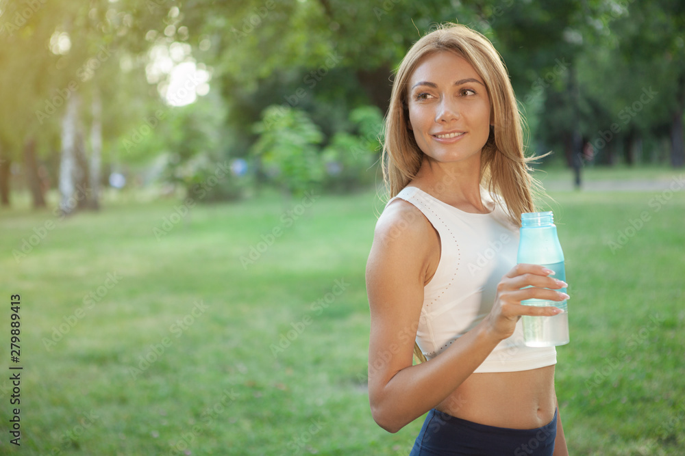 Happy healthy red haired woman smiling, looking at the copy space, holding bottle of water. Cheerful athletic woman drinking water after working out at the park Happy healthy red haired woman smiling, looking at the copy space, holding bottle of water. Cheerful athletic woman drinking water after working out at the park
