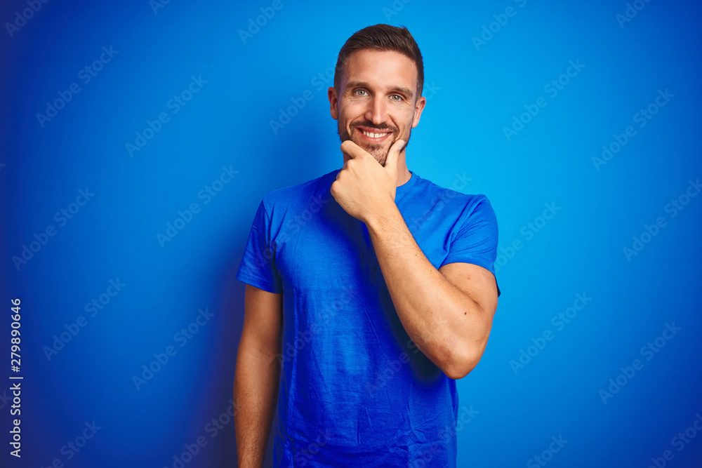 Young handsome man wearing casual t-shirt over blue isolated background looking confident at the camera smiling with crossed arms and hand raised on chin. Thinking positive. Young handsome man wearing casual t-shirt over blue isolated background looking confident at the camera smiling with crossed arms and hand raised on chin. Thinking positive.