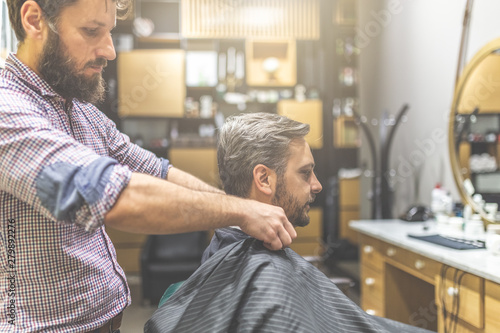 Professional barber ready to work and covering with cape bearded man on barbershop background.