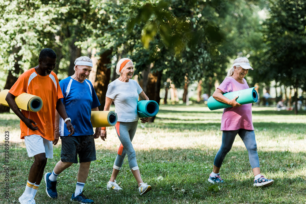 selective focus of happy multicultural and senior pensioners walking with fitness mats in park