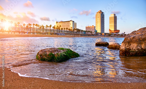 Barcelona, Spain. Panoramic view at Barceloneta beach from sea. View with stones. Sunset landscape with blue sky. Sandy coastline with palms and waves of surf. Popular touristic vacation destination. photo