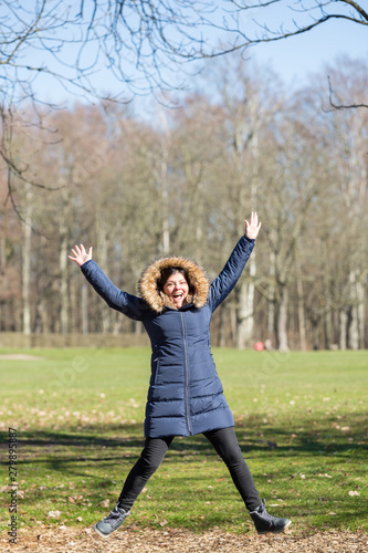 Lady jumps of joy for success. She is out in the park wearing jacket for cold weather.
