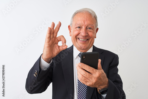 Senior grey-haired businessman using smartphone standing over isolated white background doing ok sign with fingers, excellent symbol © Krakenimages.com