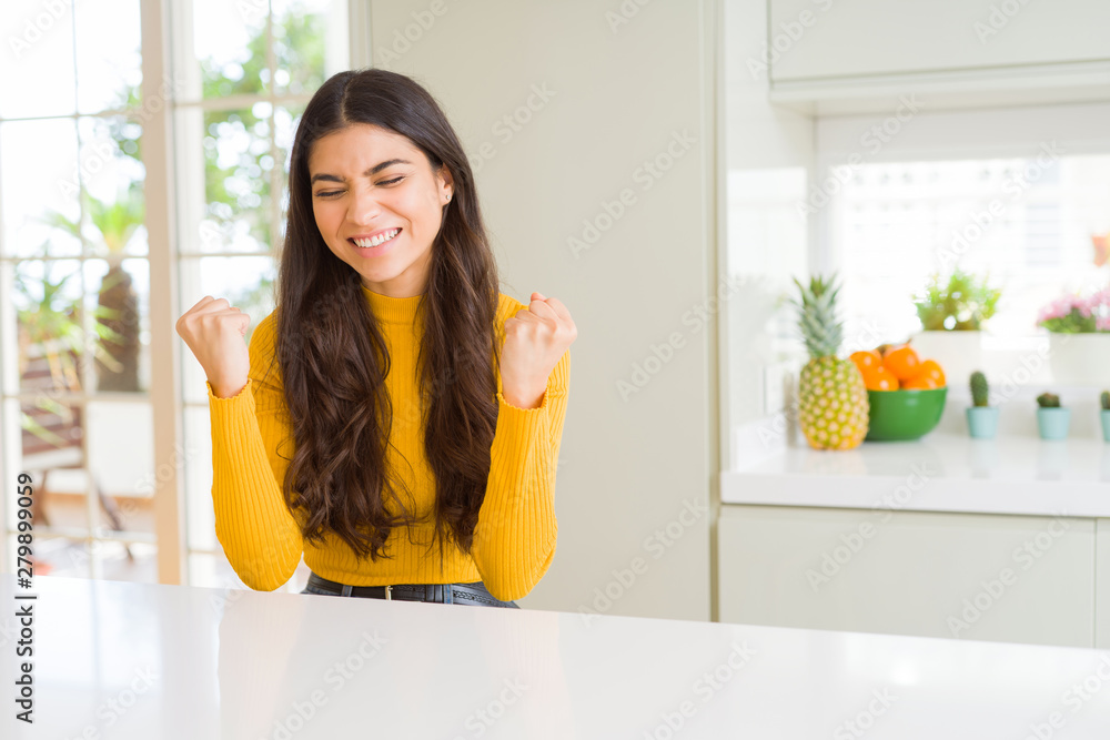 Young beautiful woman at home on white table very happy and excited doing winner gesture with arms raised, smiling and screaming for success. Celebration concept.