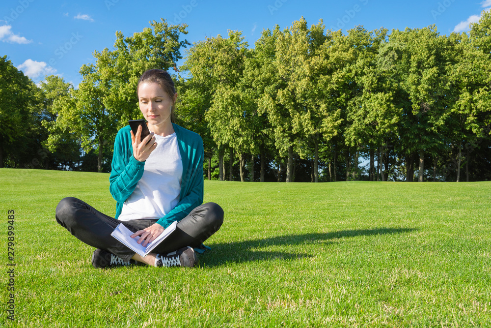 A young woman reading a message on the phone in the park on the grass. Education and knowledge.