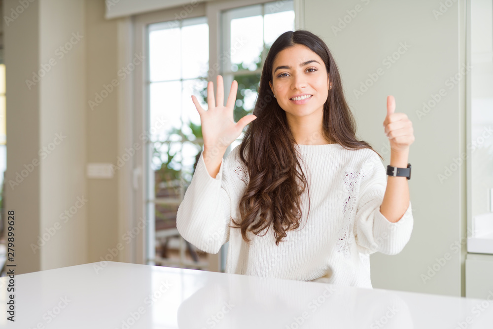 Young beautiful woman at home on white table showing and pointing up with fingers number six while smiling confident and happy. Young beautiful woman at home on white table showing and pointing up with fingers number six while smiling confident and happy.