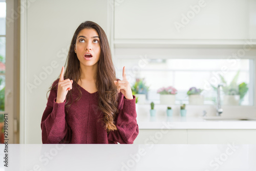 Young beautiful woman at home amazed and surprised looking up and pointing with fingers and raised arms.
