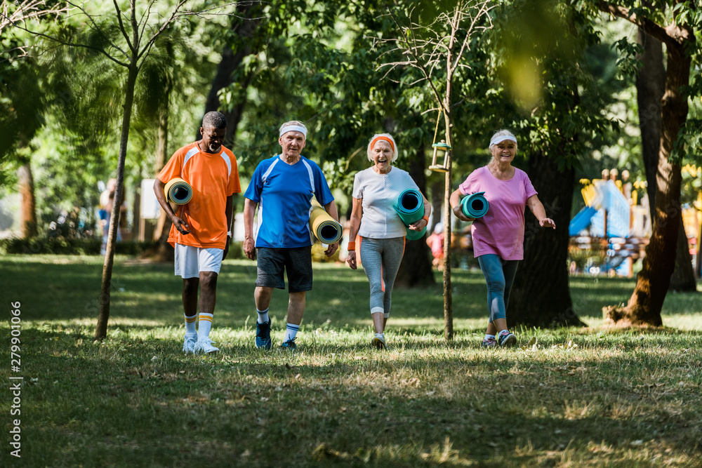 Obraz premium selective focus of cheerful multicultural and senior pensioners walking with fitness mats in park