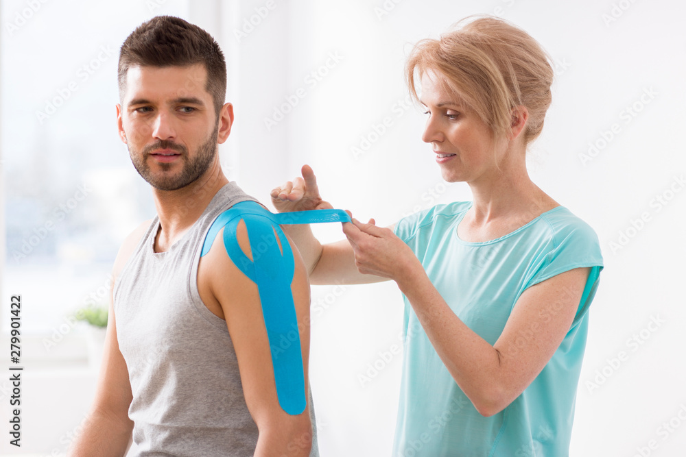 Physiotherapist covering selected fragments of young man's body with special structure patches during kinesiotaping therapy Physiotherapist covering selected fragments of young man's body with special structure patches during kinesiotaping therapy