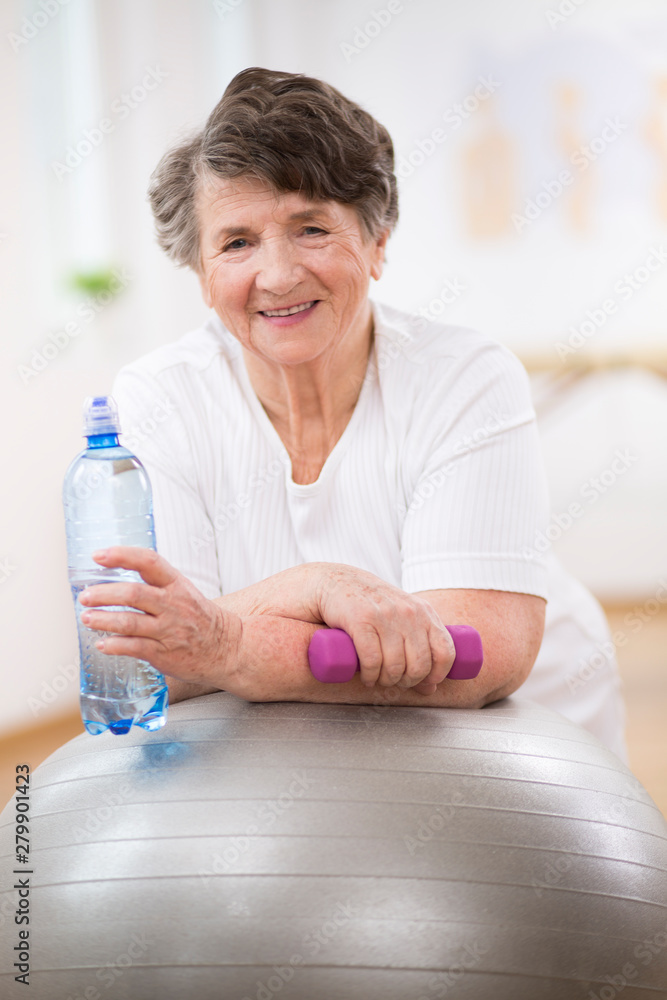 Senior woman with water bottle and dumbbell leaning on grey gymnastic ball Senior woman with water bottle and dumbbell leaning on grey gymnastic ball