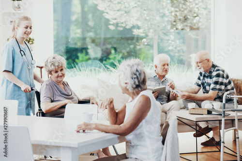 Smiling nurse helping senior woman in the wheelchair near happy elderly people