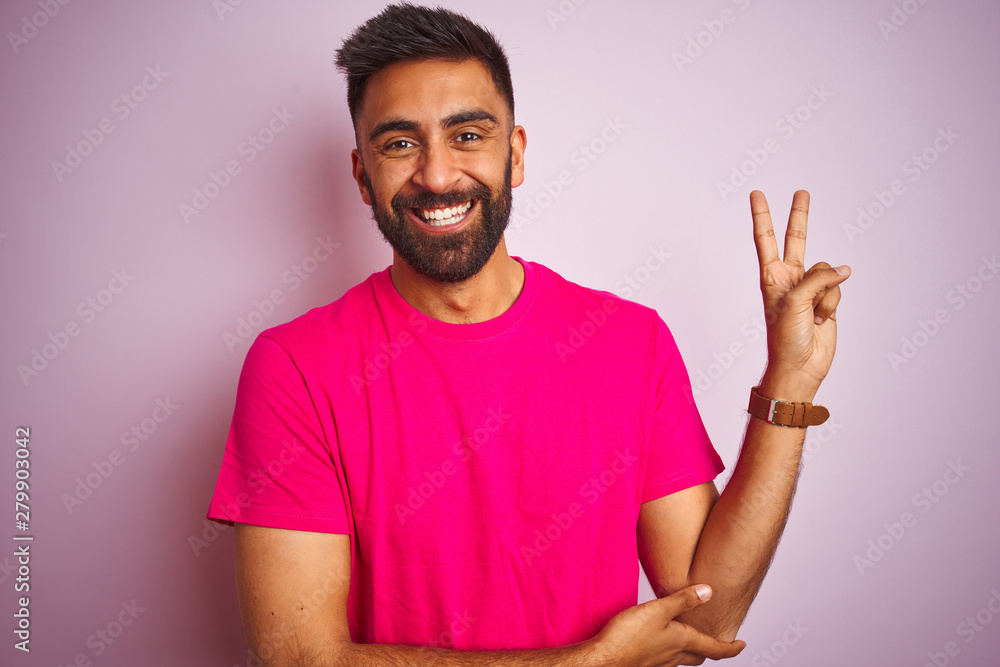 Young indian man wearing t-shirt standing over isolated pink background smiling with happy face winking at the camera doing victory sign. Number two. Young indian man wearing t-shirt standing over isolated pink background smiling with happy face winking at the camera doing victory sign. Number two.