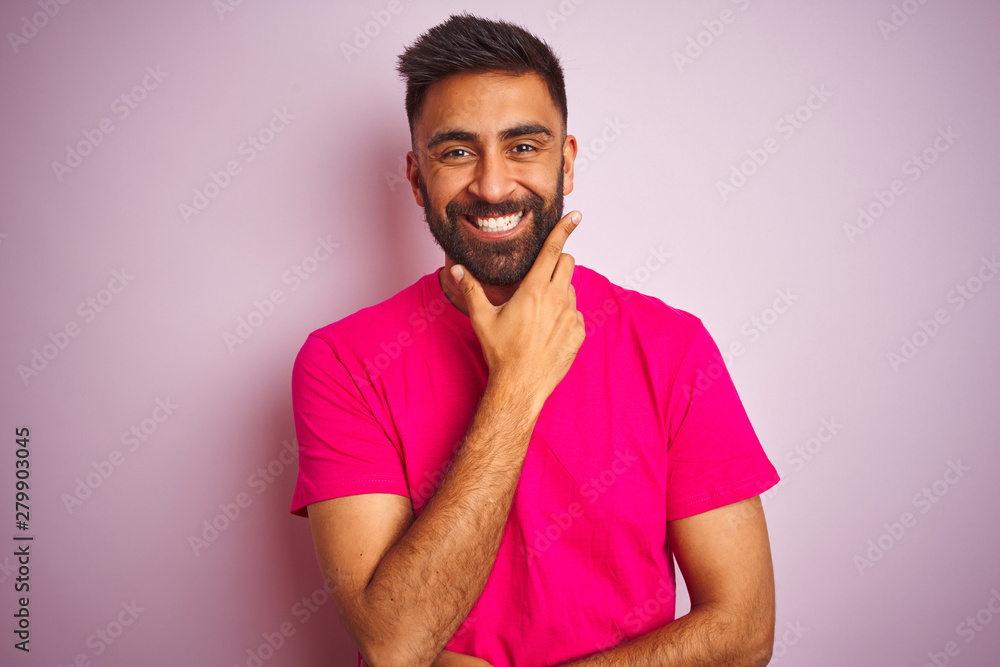 Young indian man wearing t-shirt standing over isolated pink background looking confident at the camera smiling with crossed arms and hand raised on chin. Thinking positive. Young indian man wearing t-shirt standing over isolated pink background looking confident at the camera smiling with crossed arms and hand raised on chin. Thinking positive.