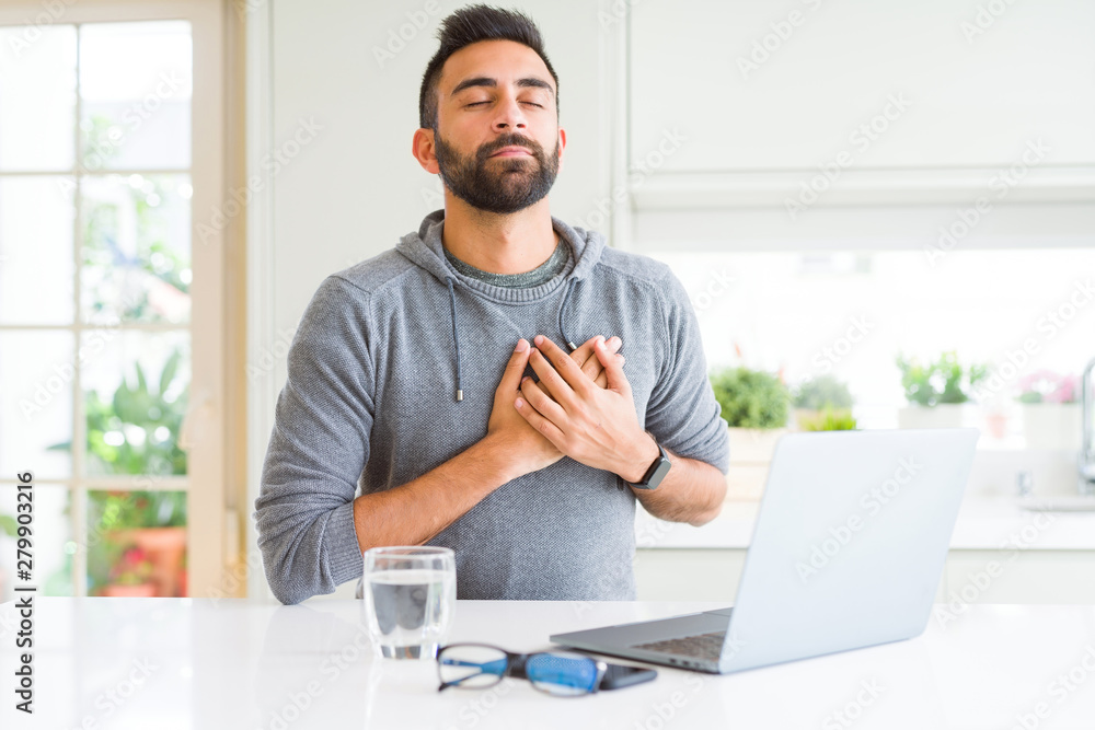 Fototapeta premium Handsome hispanic man working using computer laptop smiling with hands on chest with closed eyes and grateful gesture on face. Health concept.