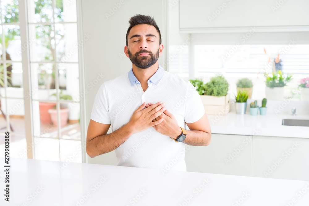 Handsome hispanic man casual white t-shirt at home smiling with hands on chest with closed eyes and grateful gesture on face. Health concept.