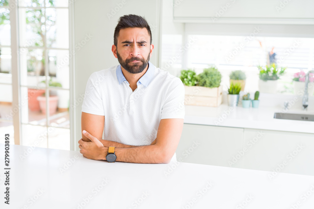 Handsome hispanic man casual white t-shirt at home skeptic and nervous, disapproving expression on face with crossed arms. Negative person.