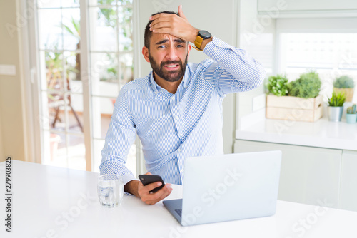 Handsome hispanic business man using laptop and smartphone stressed with hand on head, shocked with shame and surprise face, angry and frustrated. Fear and upset for mistake.