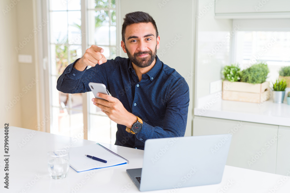 Handsome hispanic business man using smartphone and laptop at the office pointing with finger to the camera and to you, hand sign, positive and confident gesture from the front Handsome hispanic business man using smartphone and laptop at the office pointing with finger to the camera and to you, hand sign, positive and confident gesture from the front