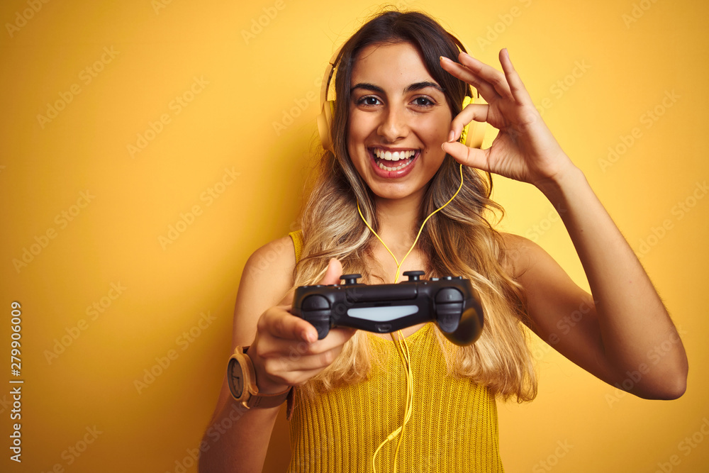 Young beautiful woman playing video game using gamepad over yellow isolated background doing ok sign with fingers, excellent symbol Young beautiful woman playing video game using gamepad over yellow isolated background doing ok sign with fingers, excellent symbol