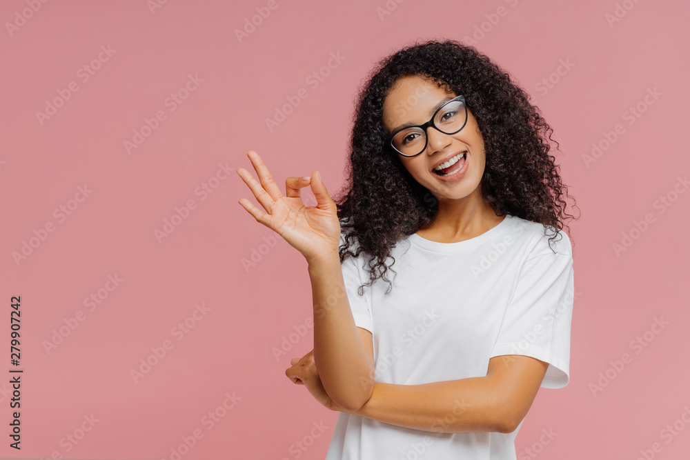 Fototapeta premium Optimistic lovely dark skinned woman makes okay gesture, tilts head, demonstrates approval, agrees with something, has happy expression, wears glasses and casual white t shirt, isolated on pink wall