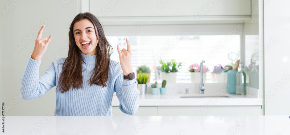 Wide angle picture of beautiful young woman sitting on white table at home shouting with crazy expression doing rock symbol with hands up. Music star. Heavy concept. Wide angle picture of beautiful young woman sitting on white table at home shouting with crazy expression doing rock symbol with hands up. Music star. Heavy concept.