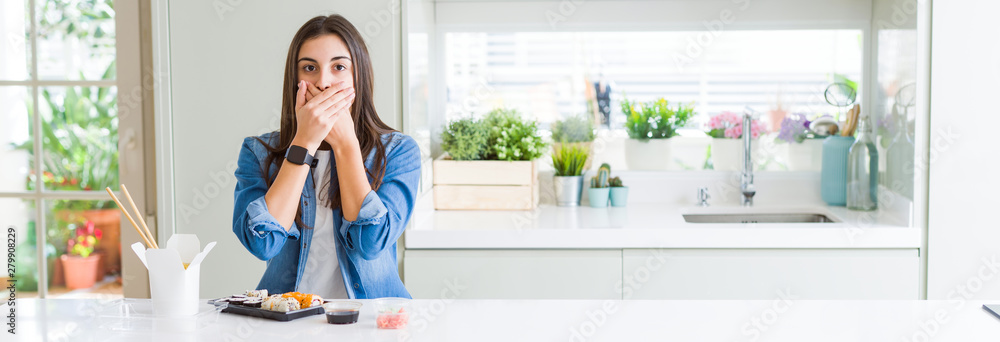 Wide angle picture of beautiful young woman eating delivery sushi shocked covering mouth with hands for mistake. Secret concept. Wide angle picture of beautiful young woman eating delivery sushi shocked covering mouth with hands for mistake. Secret concept.