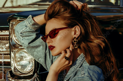 Outdoor close up fashion portrait of young beautiful fashionable woman with long luxury hair, wearing red cat eye sunglasses, denim jacket, leopard print earrings, posing near retro car, at the sunset