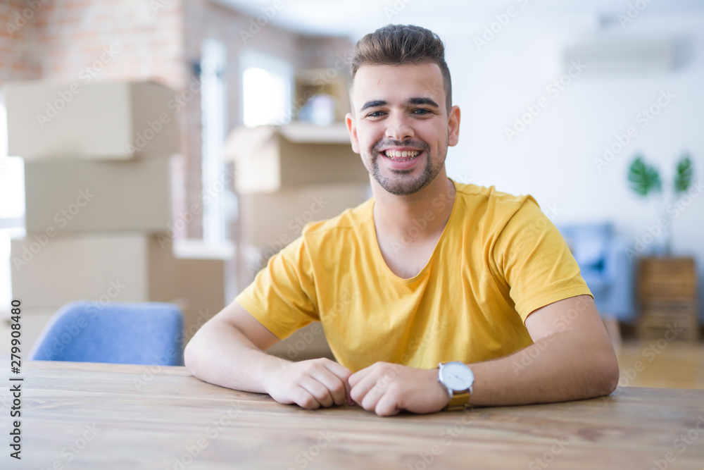 Fototapeta premium Young man sitting on the table with cardboard boxes behind him moving to new home with a happy and cool smile on face. Lucky person.