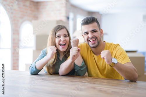 Young couple sitting on the table movinto to new home with carboard boxes behind them very happy and excited doing winner gesture with arms raised, smiling and screaming for success. 