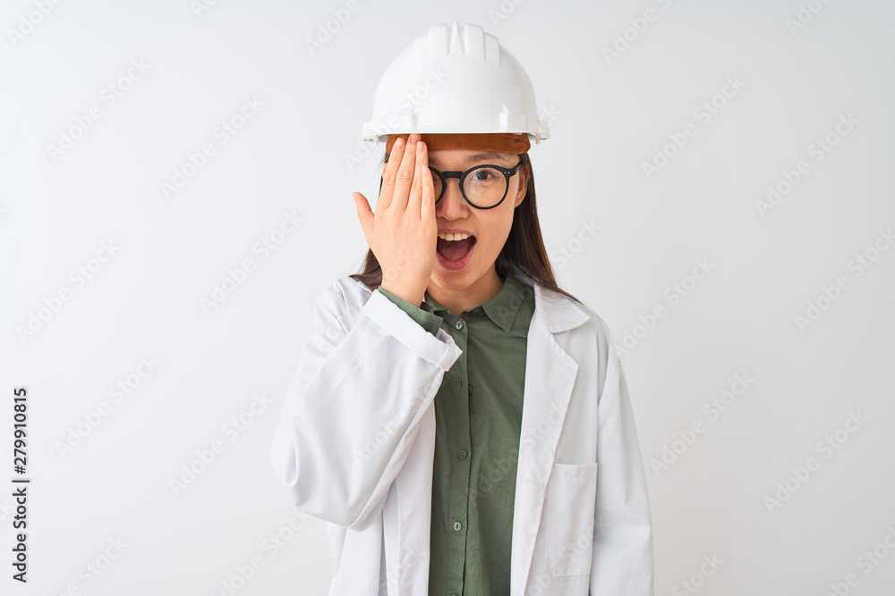 Young chinese engineer woman wearing coat helmet glasses over isolated white background covering one eye with hand, confident smile on face and surprise emotion. Young chinese engineer woman wearing coat helmet glasses over isolated white background covering one eye with hand, confident smile on face and surprise emotion.