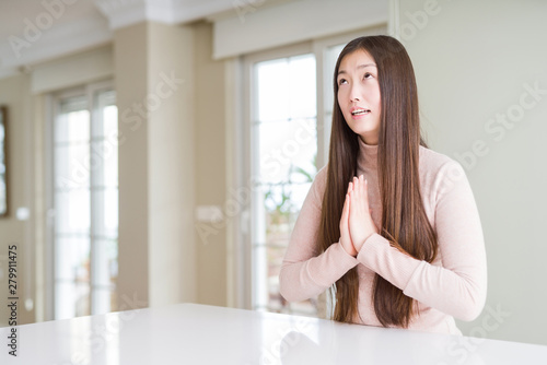 Beautiful Asian woman wearing casual sweater on white table begging and praying with hands together with hope expression on face very emotional and worried. Asking for forgiveness. Religion concept.