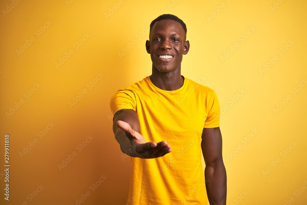Young african american man wearing casual t-shirt standing over isolated yellow background smiling cheerful offering palm hand giving assistance and acceptance. Young african american man wearing casual t-shirt standing over isolated yellow background smiling cheerful offering palm hand giving assistance and acceptance.
