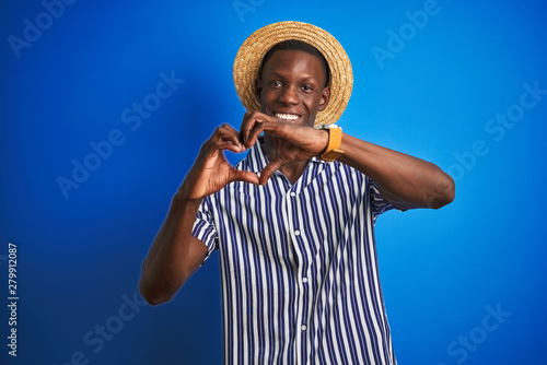 African american man wearing striped shirt and summer hat over isolated blue background smiling in love showing heart symbol and shape with hands. Romantic concept.