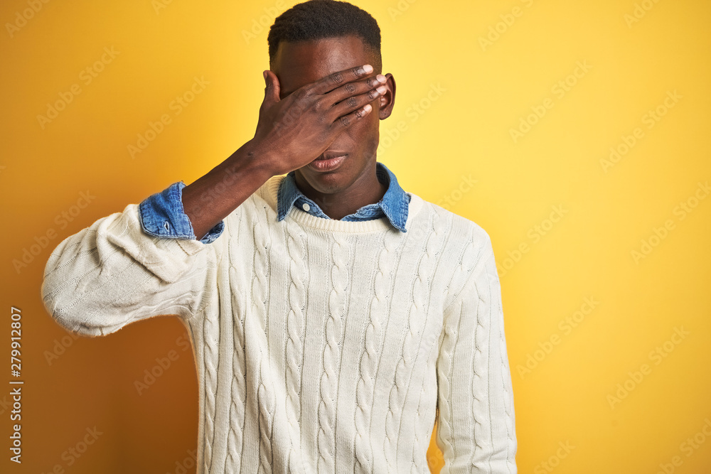 African american man wearing denim shirt and white sweater over isolated yellow background covering eyes with hand, looking serious and sad. Sightless, hiding and rejection concept African american man wearing denim shirt and white sweater over isolated yellow background covering eyes with hand, looking serious and sad. Sightless, hiding and rejection concept