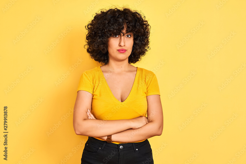 Young arab woman with curly hair wearing t-shirt standing over isolated yellow background skeptic and nervous, disapproving expression on face with crossed arms. Negative person. Young arab woman with curly hair wearing t-shirt standing over isolated yellow background skeptic and nervous, disapproving expression on face with crossed arms. Negative person.