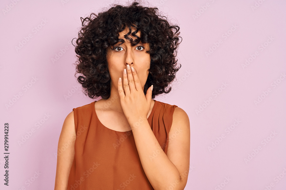 Young arab woman with curly hair wearing t-shirt over isolated pink background cover mouth with hand shocked with shame for mistake, expression of fear, scared in silence, secret concept Young arab woman with curly hair wearing t-shirt over isolated pink background cover mouth with hand shocked with shame for mistake, expression of fear, scared in silence, secret concept