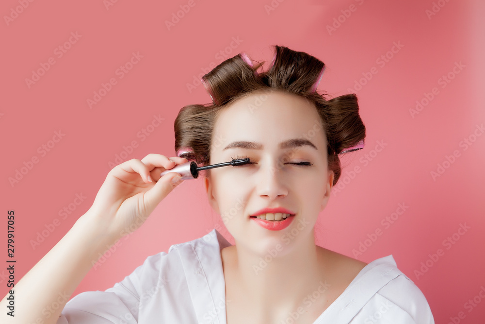 Fototapeta premium nice cheerful young the girl in the curler paints an eyebrow on pink background