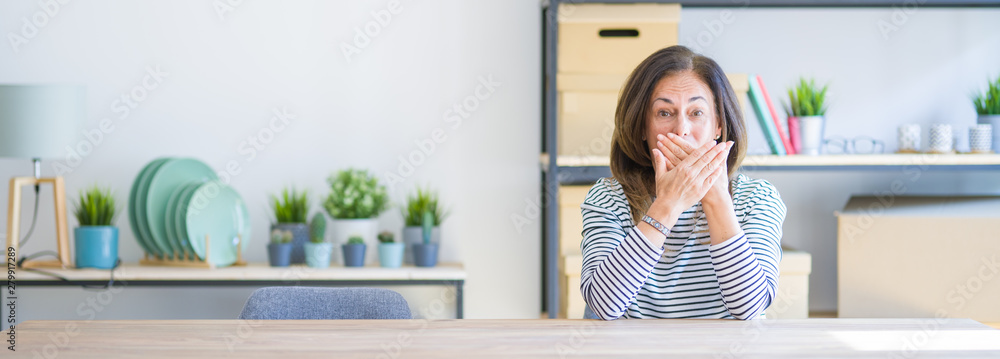Wide angle photo of middle age senior woman sitting at the table at home shocked covering mouth with hands for mistake. Secret concept. Wide angle photo of middle age senior woman sitting at the table at home shocked covering mouth with hands for mistake. Secret concept.