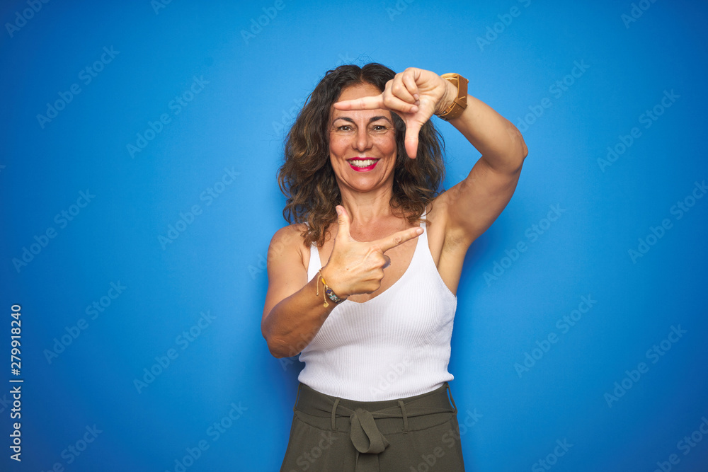 Middle age senior woman with curly hair standing over blue isolated background smiling making frame with hands and fingers with happy face. Creativity and photography concept.