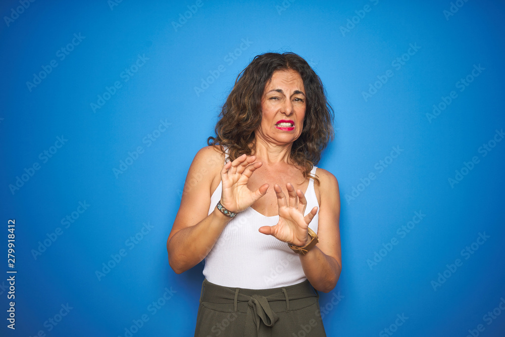 Middle age senior woman with curly hair standing over blue isolated background disgusted expression, displeased and fearful doing disgust face because aversion reaction. With hands raised. Middle age senior woman with curly hair standing over blue isolated background disgusted expression, displeased and fearful doing disgust face because aversion reaction. With hands raised.