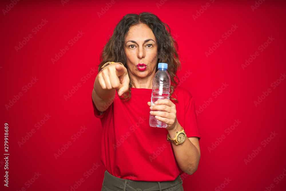 Middle age senior woman holding plastic water bottle over red isolated background pointing with finger to the camera and to you, hand sign, positive and confident gesture from the front Middle age senior woman holding plastic water bottle over red isolated background pointing with finger to the camera and to you, hand sign, positive and confident gesture from the front