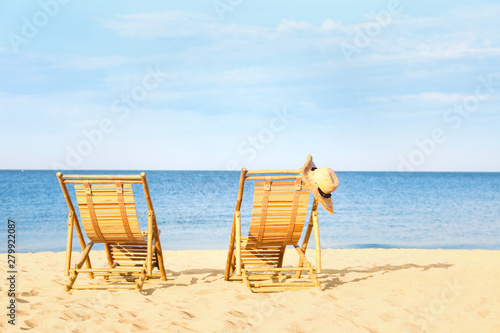 Empty wooden sunbeds with hat on sandy shore. Beach accessories