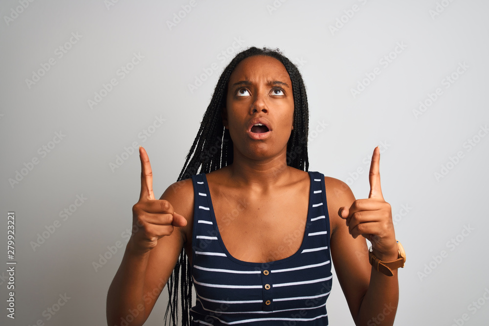 Young african american woman wearing striped t-shirt standing over isolated white background amazed and surprised looking up and pointing with fingers and raised arms. Young african american woman wearing striped t-shirt standing over isolated white background amazed and surprised looking up and pointing with fingers and raised arms.