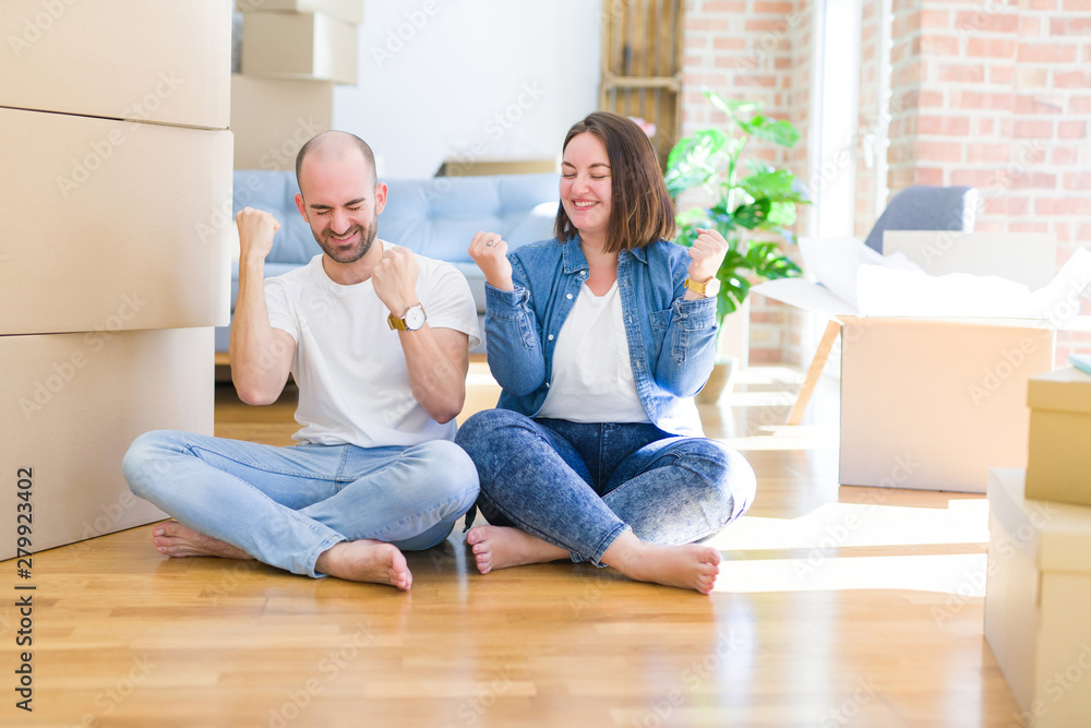 Young couple sitting on the floor arround cardboard boxes moving to a new house very happy and excited doing winner gesture with arms raised, smiling and screaming for success. Celebration concept.