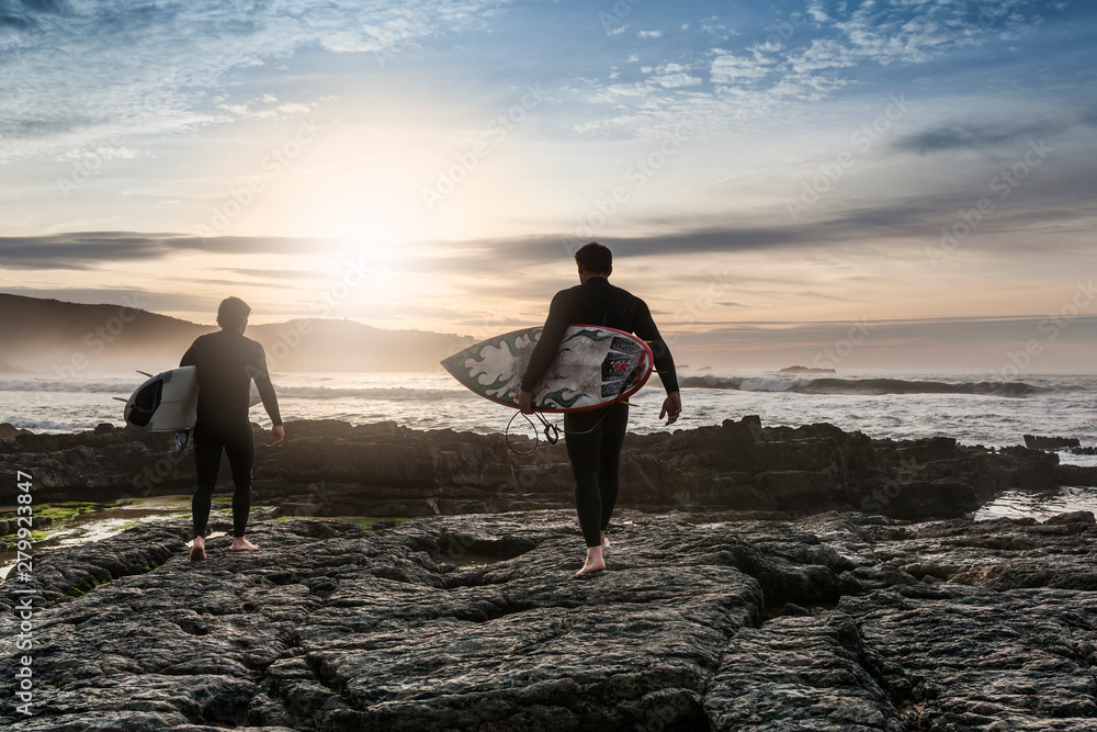 Fototapeta premium two male surfers on rocks with their surfboard on their way to get into the water to surf at sunset