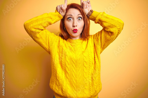 Beautiful redhead woman wearing winter sweater standing over isolated yellow background doing funny gesture with finger over head as bull horns © Krakenimages.com