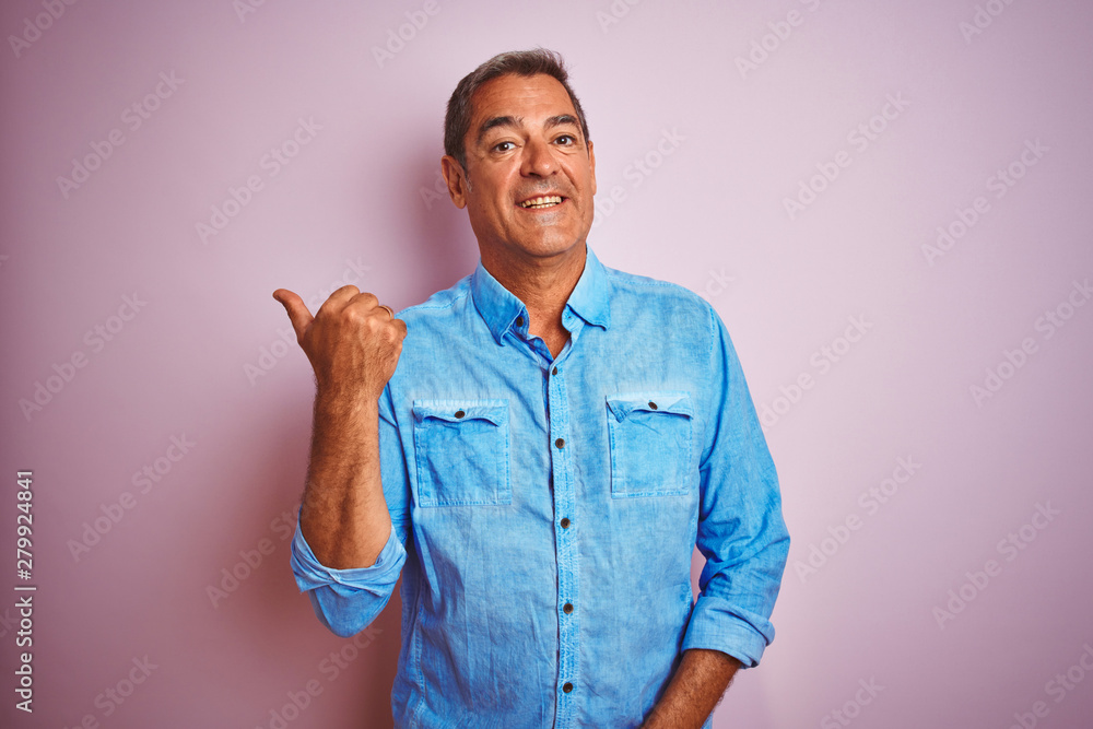 Handsome middle age man wearing blue denim shirt standing over isolated pink background smiling with happy face looking and pointing to the side with thumb up. Handsome middle age man wearing blue denim shirt standing over isolated pink background smiling with happy face looking and pointing to the side with thumb up.