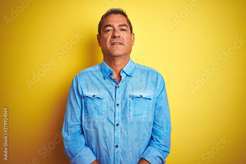 Handsome middle age man wearing denim shirt standing over isolated yellow background with serious expression on face. Simple and natural looking at the camera. © Krakenimages.com